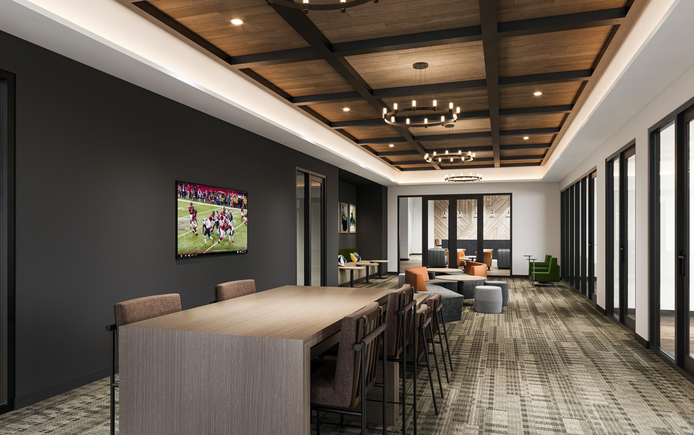 Modern lounge with wood ceiling, long table, chairs, sofas, TV on wall, and large windows in back.